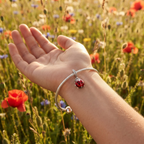 Lucky Ladybug Charm on a silver snake-chain bracelet, worn on an open hand in a field of poppies