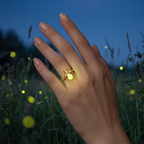 Gold Luminous Firefly Ring glowing in the dark on a hand in a summer meadow at night with fireflies